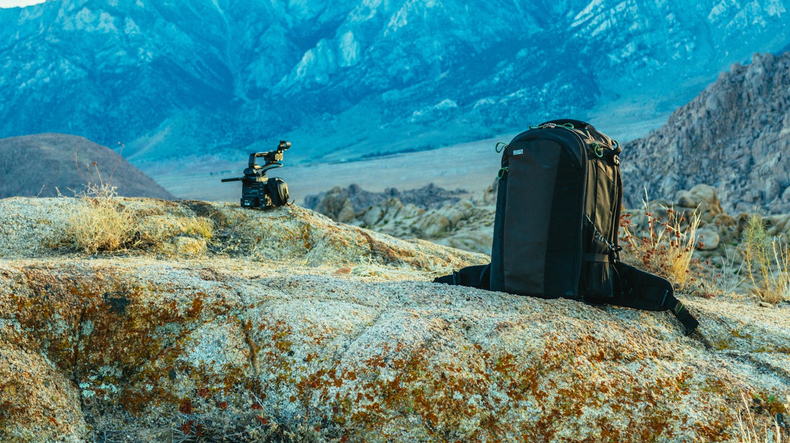 Professional camera backpack resting on rocks in the Alabama Hills with a cinema camera rig beside it and dramatic blue Sierra Nevada peaks behind — the kind of kit that demands a serious bag system