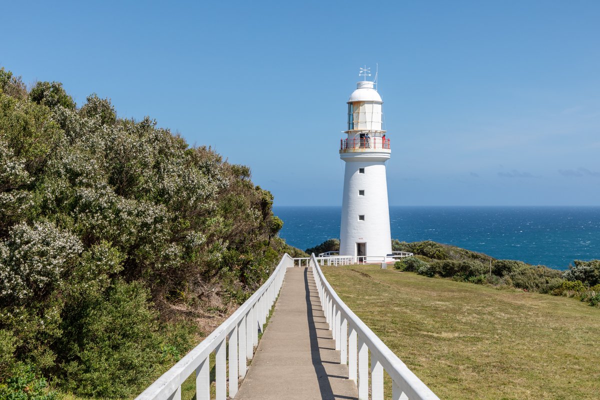 The white Cape Otway lighthouse on a grassy headland with a walkway leading toward it and the ocean behind