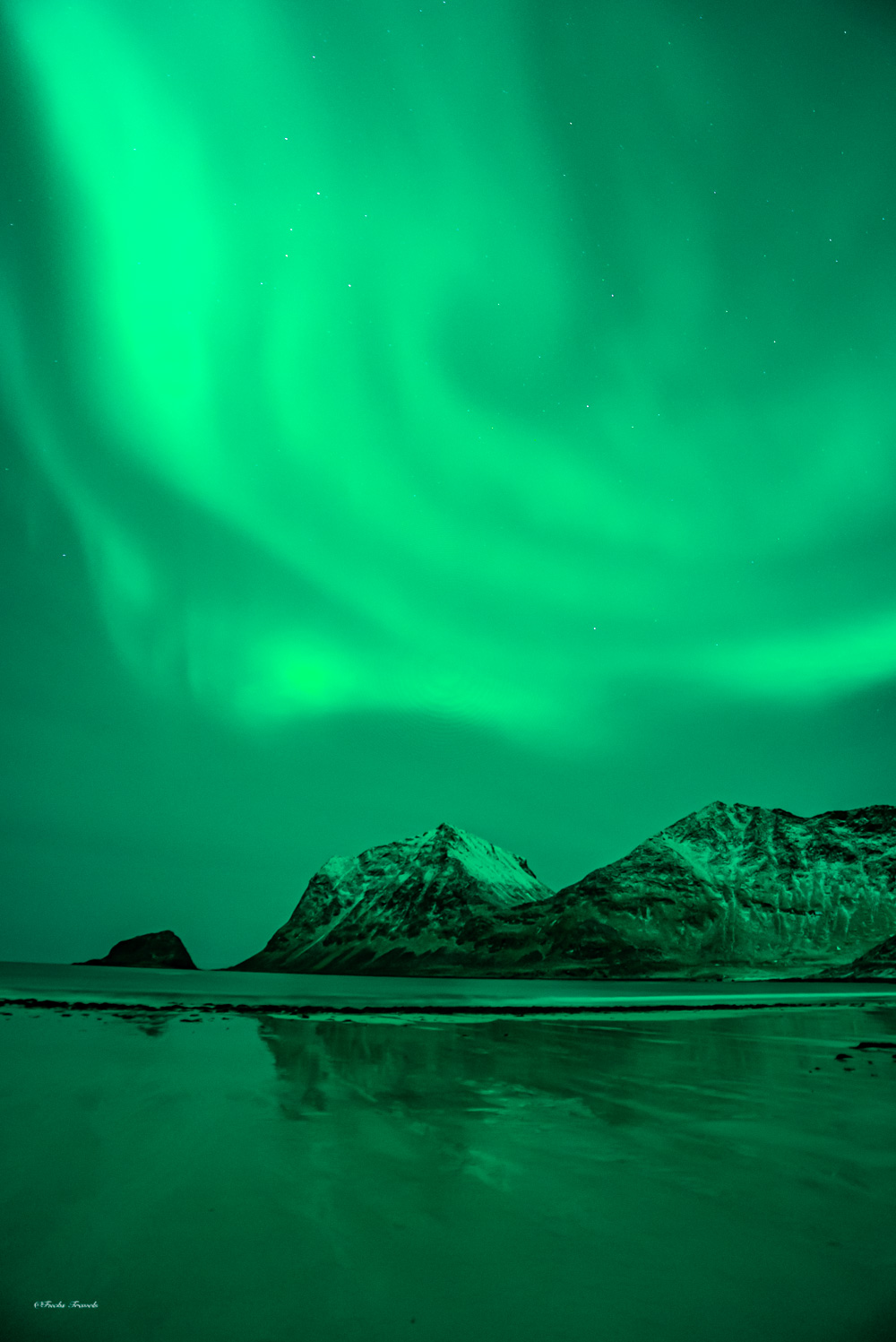 Intense green aurora borealis above snow-capped Lofoten peaks with reflections on beach water